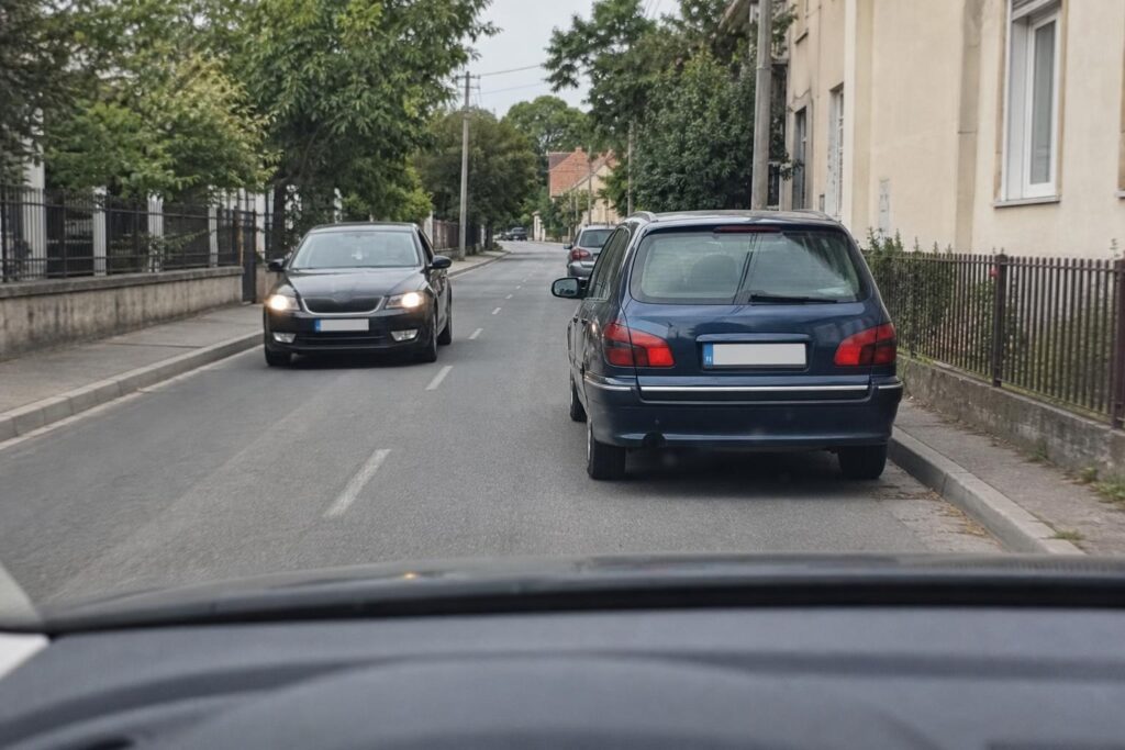 Autó akadályt kerül ki egy keskeny kétirányú úton, miközben szemből jármű érkezik