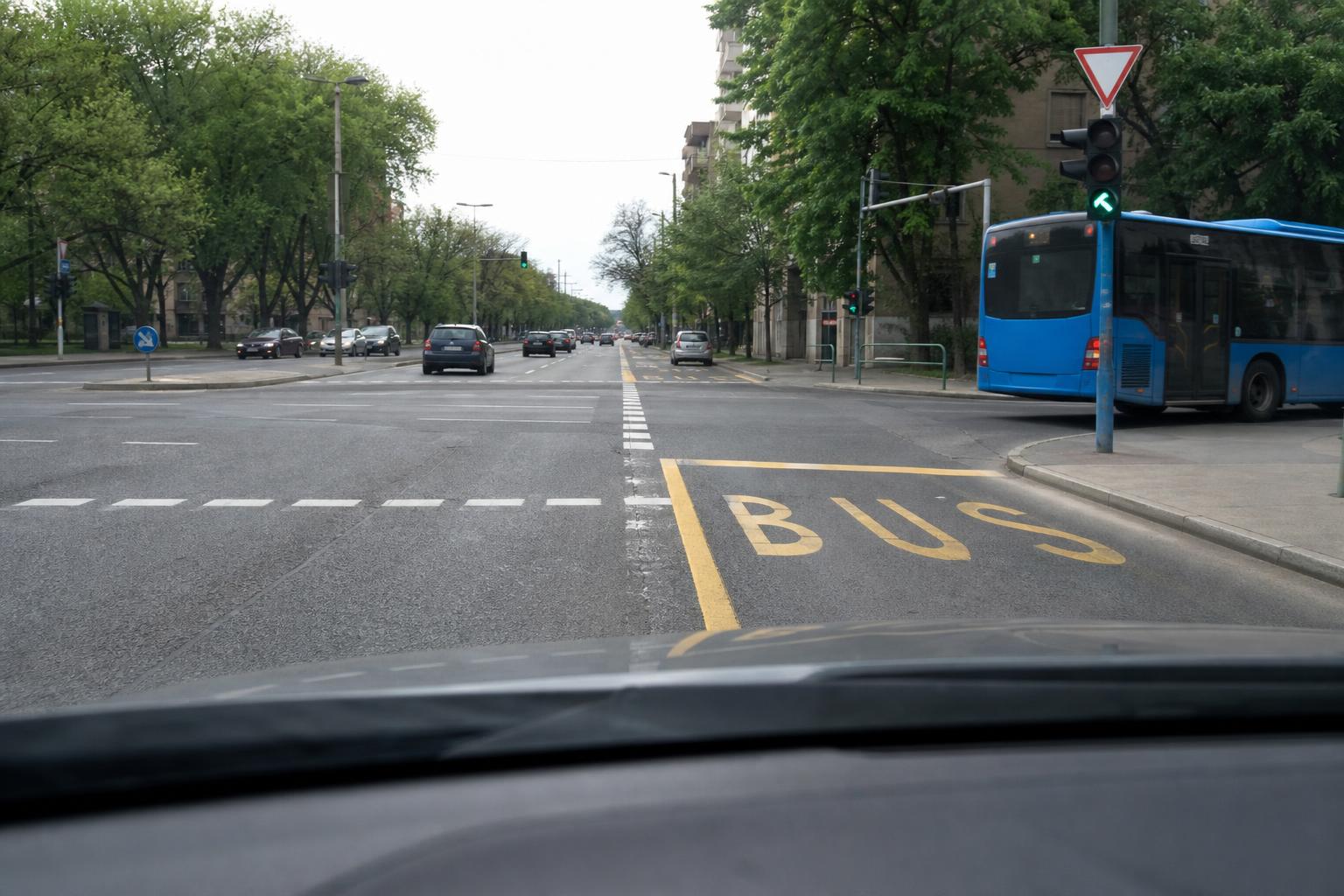 Autó jobbra kanyarodásra készül egy városi buszsáv keresztezésével, a közelben autóbusszal és jól látható útburkolati jelekkel.