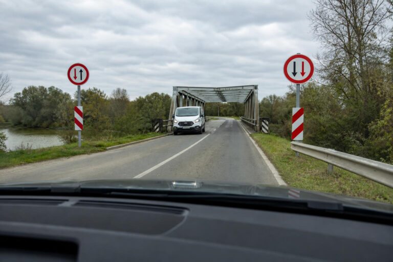 Keskeny hídra érkező autó szembejövő járművel és elsőbbséget jelző közúti táblákkal