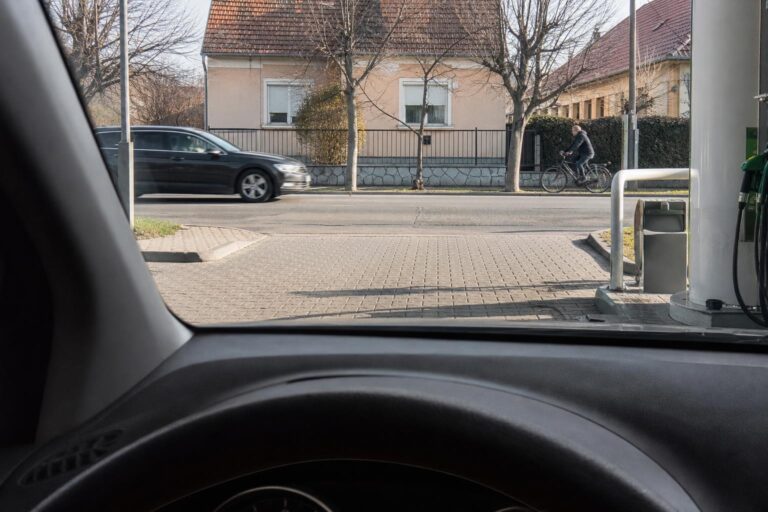 Autó várakozik benzinkút kijáratánál, miközben a járdán kerékpáros, az úton pedig jármű halad el