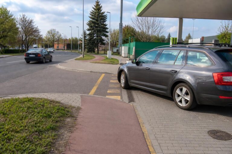 Autó hajt ki egy benzinkút kijáratán a járdán és kerékpárúton át, miközben jármű közeledik a közúton