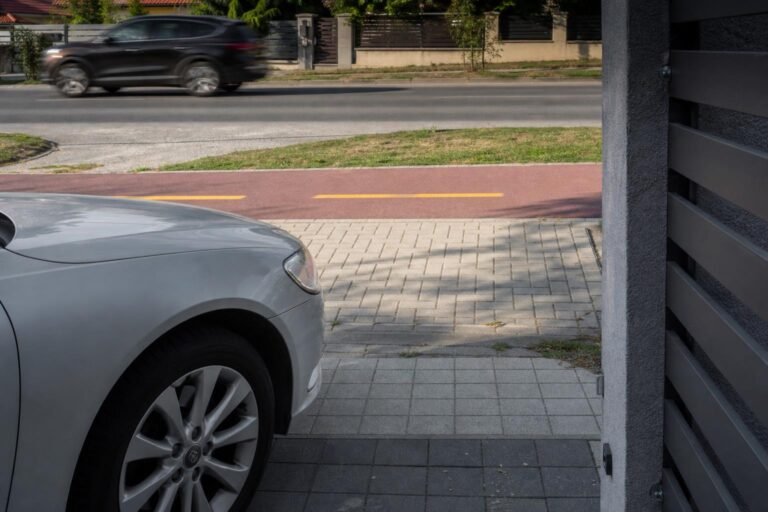 Autó óvatosan hajt ki egy kapubejáróból a járda és kerékpárút keresztezésén át