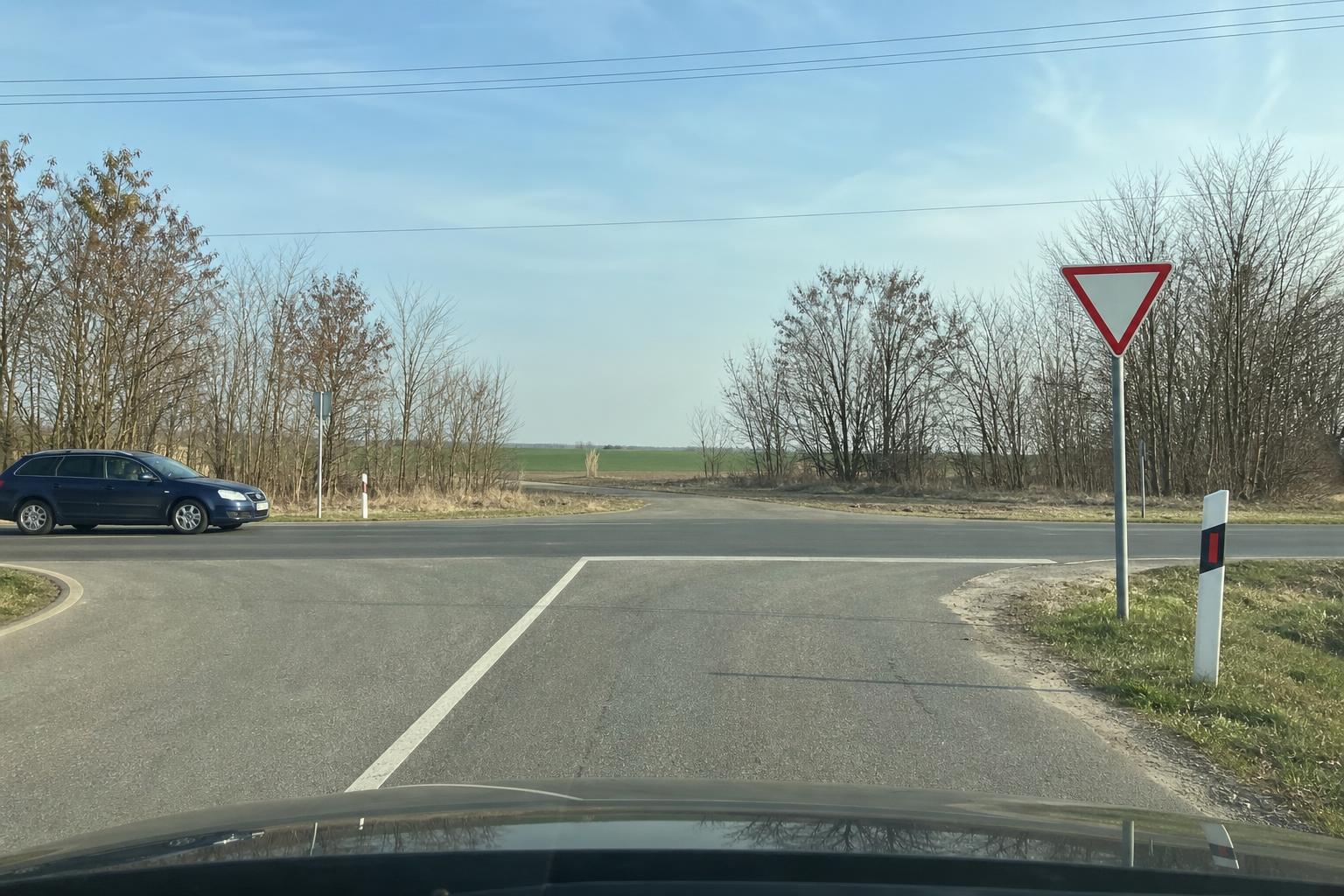 Autó közeledik egy táblákkal és útburkolati jelekkel szabályozott T alakú kereszteződéshez
