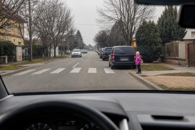 Gyermek áll a járda szélén egy iskola közelében, miközben autó közelít a gyalogos-átkelőhelyhez