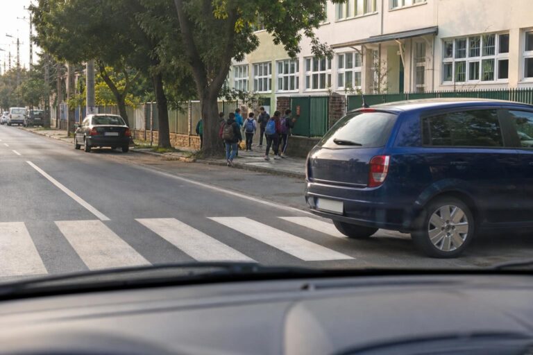 Iskolai gyalogátkelőhely előtt lassító autó, a járdán gyerekekkel és részben takaró parkoló járművel