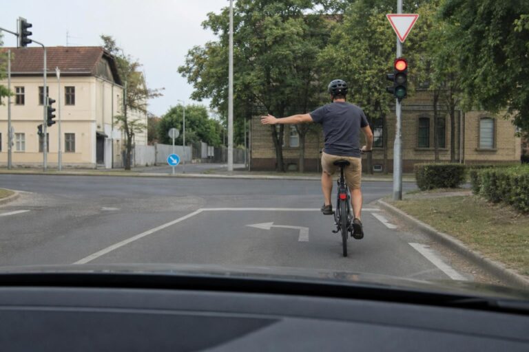 Balra kanyarodást jelző kerékpáros egy városi kereszteződésben, mögötte lassító autóval