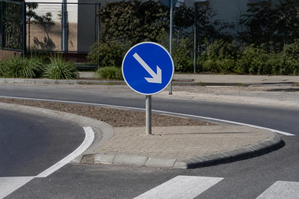 Kék, kör alakú kikerülési irányt jelző tábla egy forgalmi terelősziget mellett