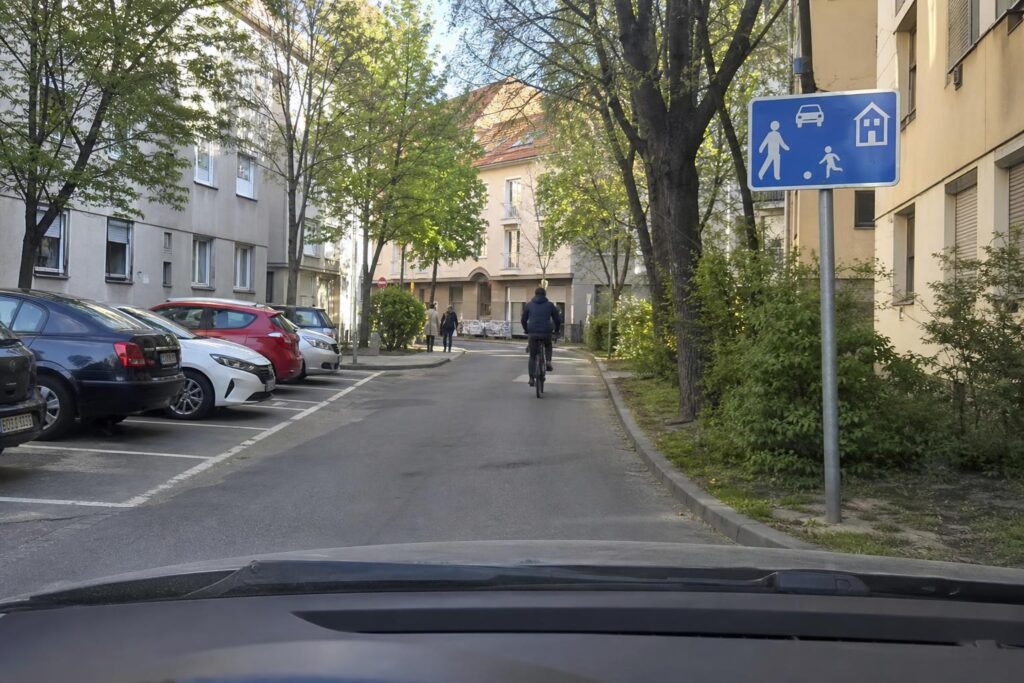 Lakó-pihenő övezetet jelző tábla csendes magyar lakóutcában, kijelölt parkolóhelyekkel és az úttesten közlekedő gyalogosokkal
