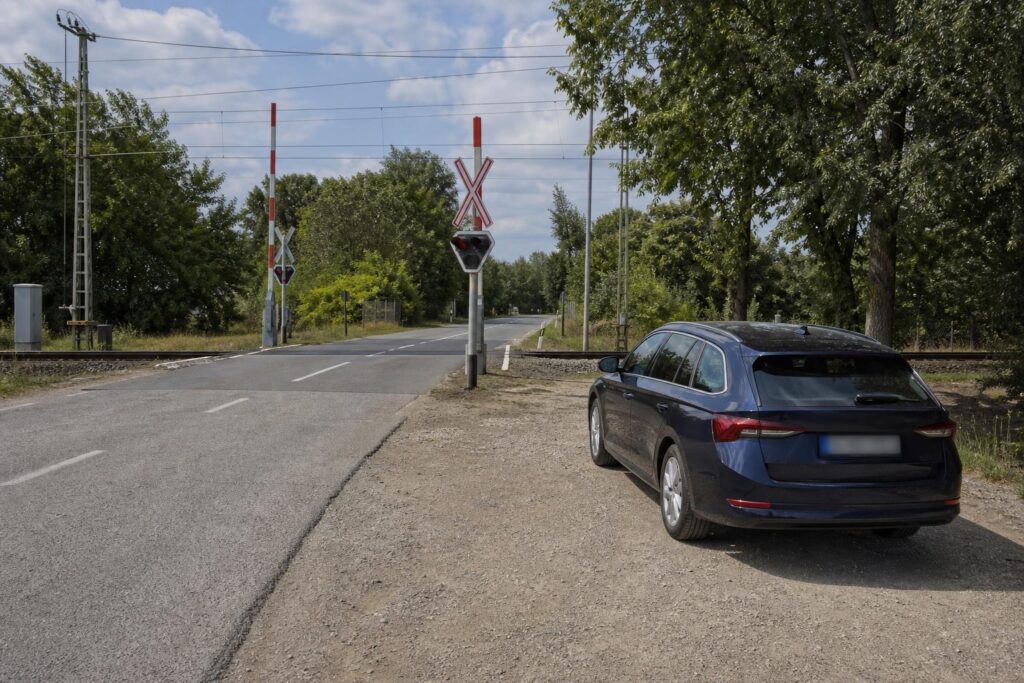 Személyautó a vasúti átjárótól biztonságos távolságban, háttérben sorompóval, vasúti jelzőfénnyel és keresztjelző táblával