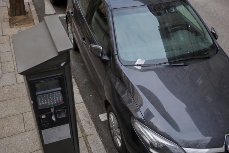 Fizetős parkolóhelyen álló autó szélvédőjén elhelyezett parkolási értesítés és a háttérben parkolóautomata