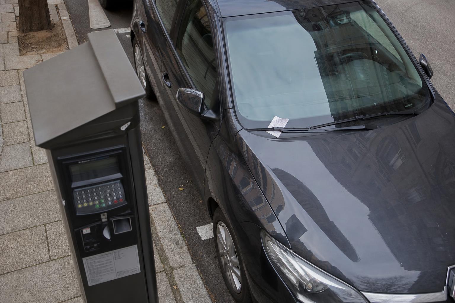 Fizetős parkolóhelyen álló autó szélvédőjén elhelyezett parkolási értesítés és a háttérben parkolóautomata