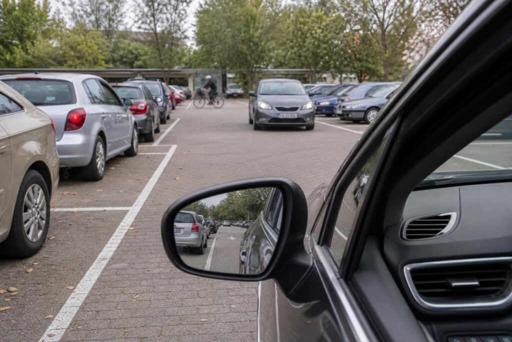 Autó kiállása kijelölt parkolóhelyről egy parkoló belső útjára, közeledő járművel és kerékpárossal