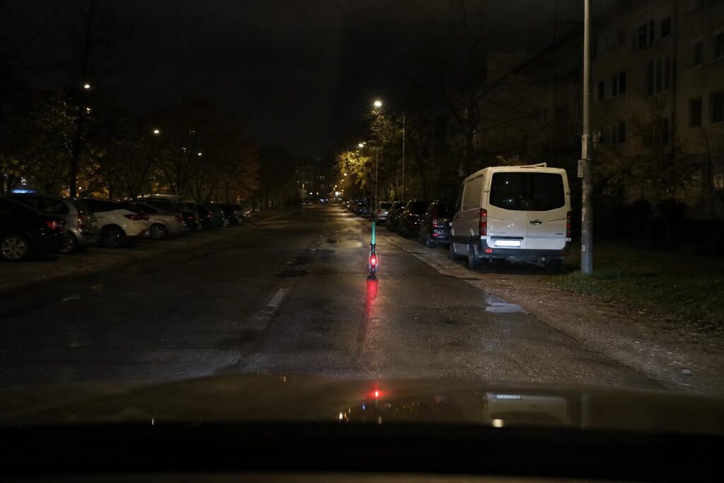Kivilágított roller halad éjszaka az úttesten nedves aszfalton, autós nézőpontjából