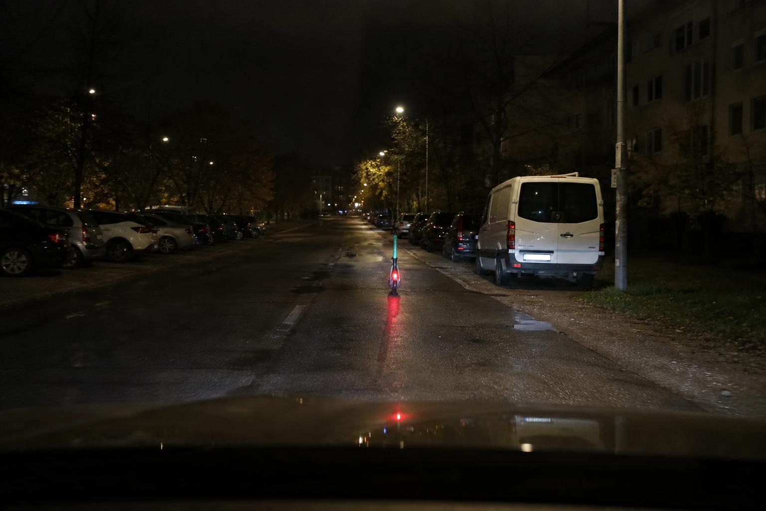 Kivilágított roller halad éjszaka az úttesten nedves aszfalton, autós nézőpontjából