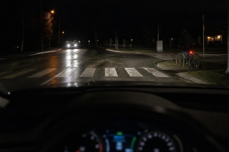 Sötét, nedves úton haladó autó fényszórója egy kereszteződés és az út szélén várakozó kerékpáros előtt