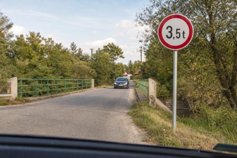 Személyautó közeledik piros szegélyű súlykorlátozó közlekedési táblához egy keskeny úton