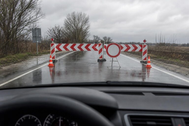 Autó előtt látható piros-fehér útzár és hivatalos útlezáró tábla egy nedves úton
