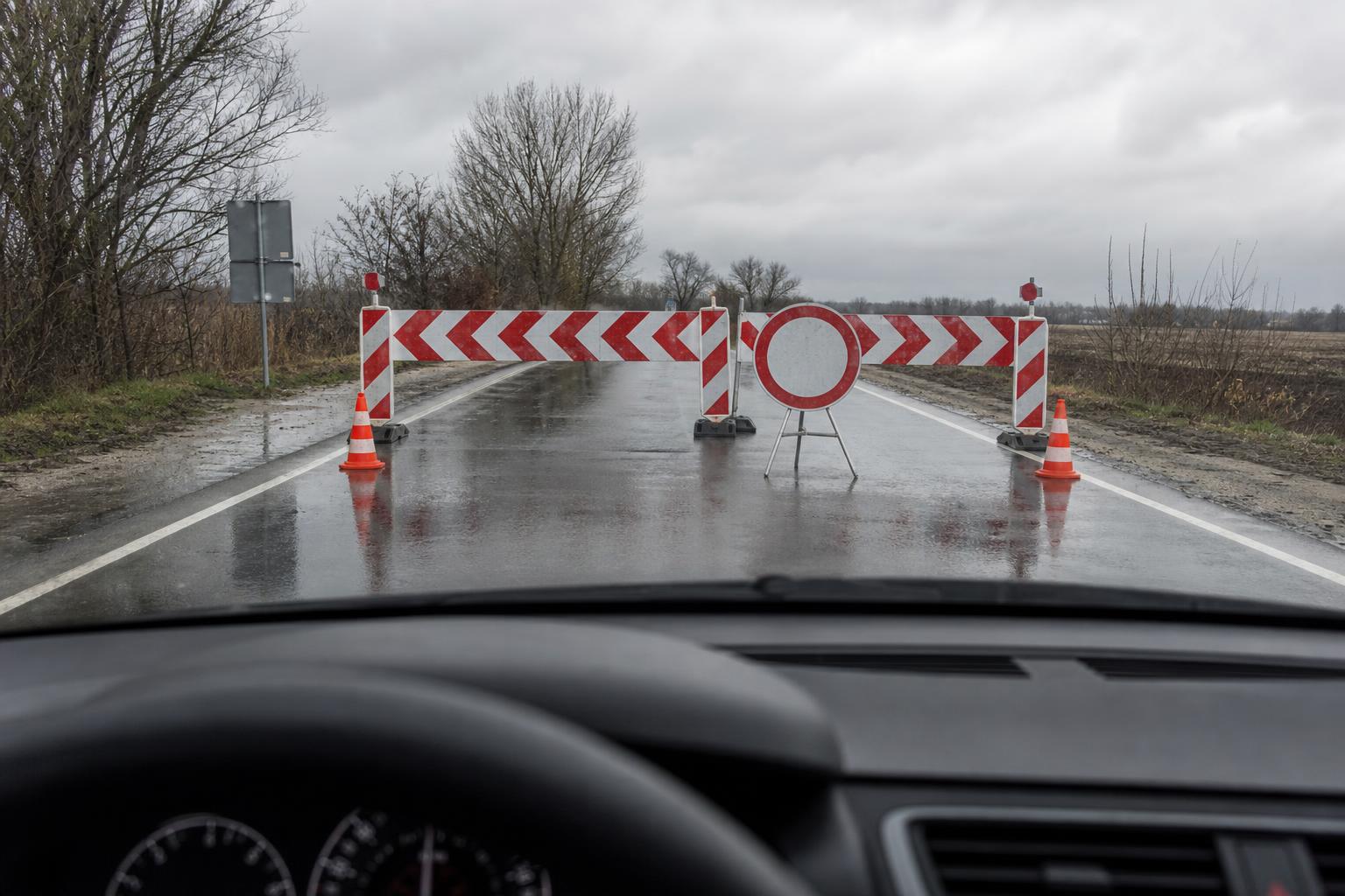 Autó előtt látható piros-fehér útzár és hivatalos útlezáró tábla egy nedves úton