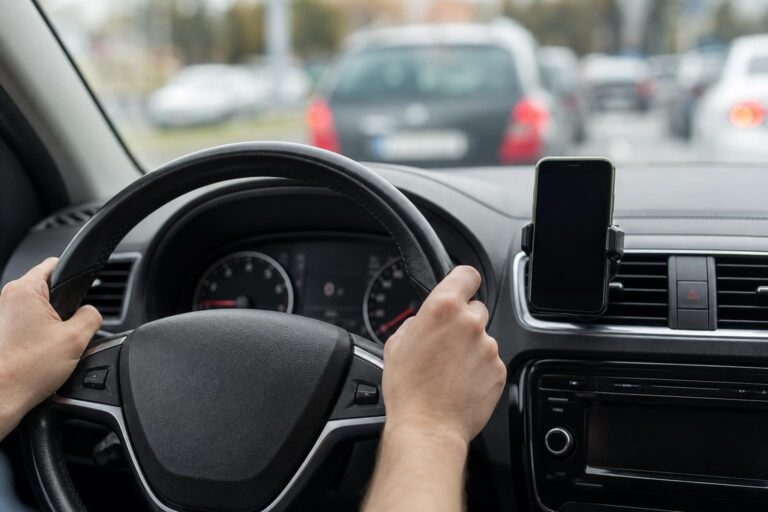 Biztonságosan rögzített, kijelzővel lefelé fordított mobiltelefon az autó műszerfalán, miközben a kéz a kormányon van