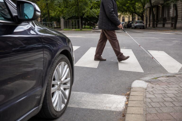 Autó áll meg a zebránál, miközben fehér bottal közlekedő vak gyalogos kel át az úton