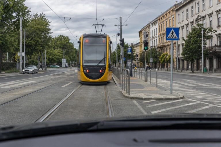 Autó halad egy városi villamos mögött, előtte villamosmegálló és gyalogossziget látható