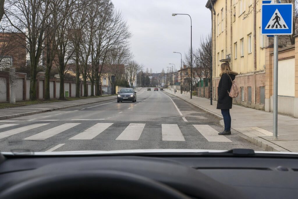 Autó lassít egy kijelölt gyalogos-átkelőhely előtt, miközben egy gyalogos a járdaszegélynél várakozik